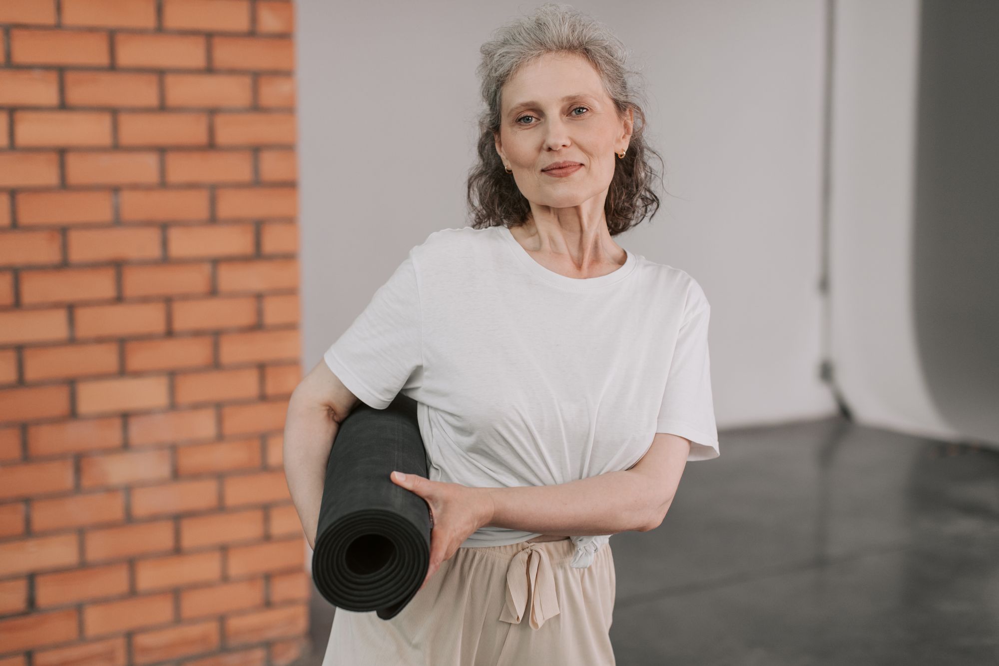 A woman seated beside a brick wall holding a rolled exercise mat