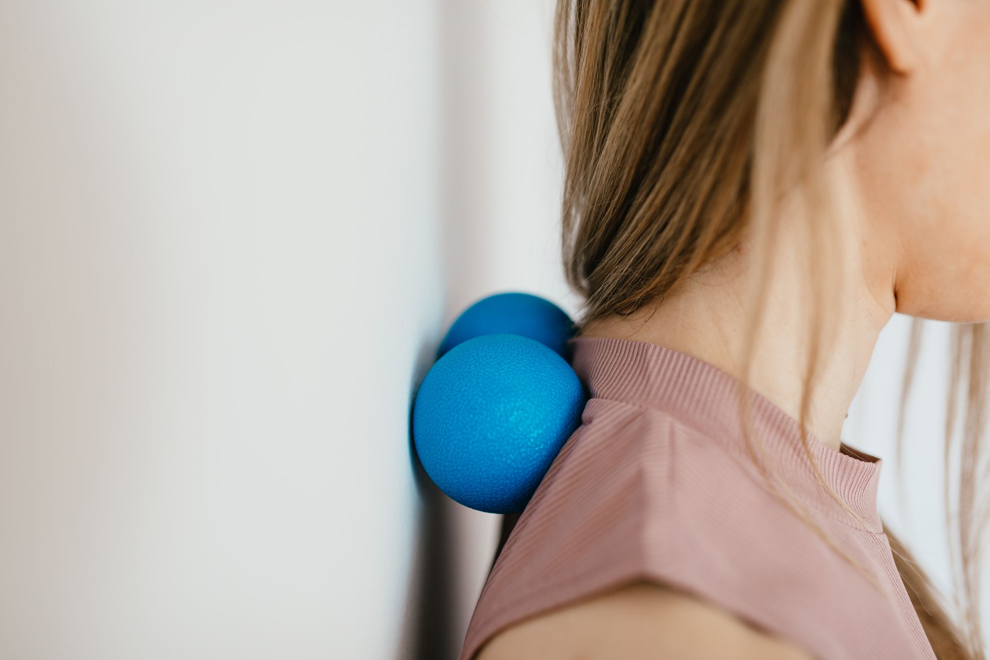 A woman uses a blue massage ball against her upper shoulder near the neck