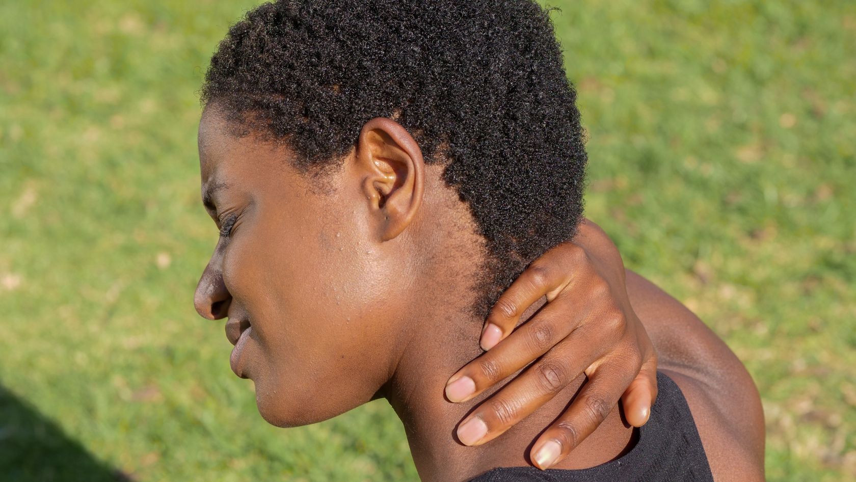 Close-up of a person holding the back of their neck outdoors against a grassy background
