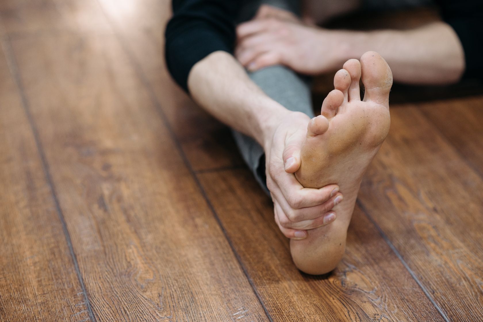 Close-up of a person holding the sole of their bare foot while seated on a wooden floor