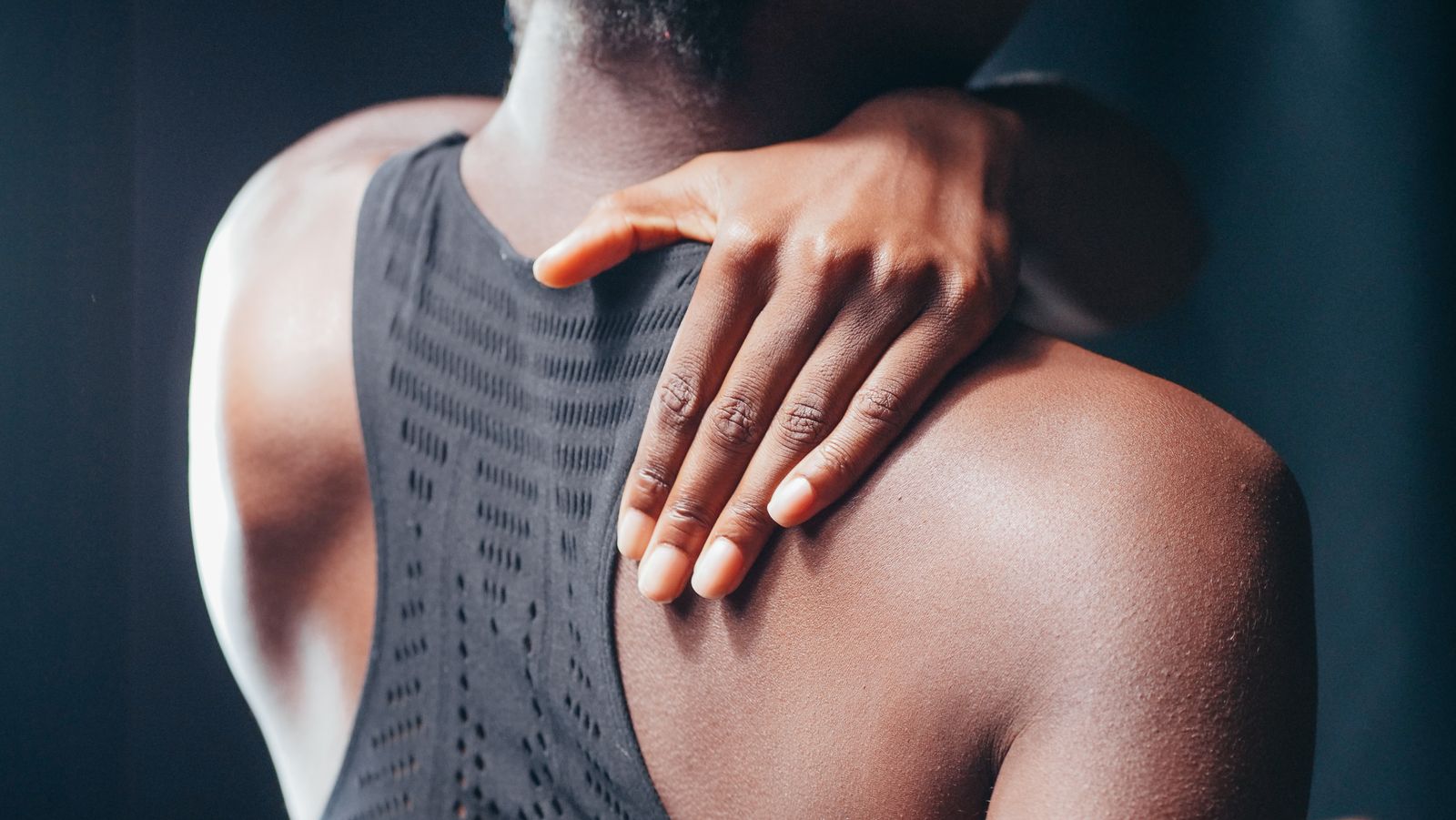 Person in a black sleeveless top reaching back to rest a hand on their upper shoulder
